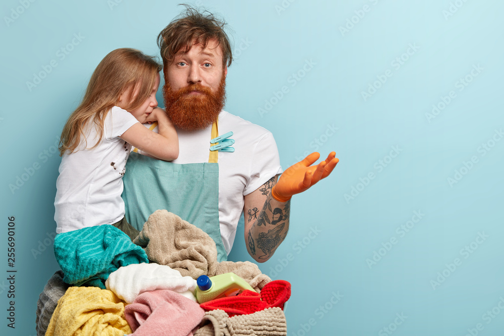 Busy overloaded father looks after little daughter and does household chores, has puzzled fatigue expression, has much work, washes clothes, spreads hand in bewilderment, isolated on blue wall - obrazy, fototapety, plakaty 