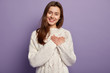 © WHstudio Leushin N - Friendly pleased woman has heart filled with love and gratitude, keeps both hands on chest, has lovely sincere smile, wears oversized jumper, stands against purple background, sympatizes friend