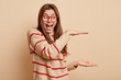 Photo of cheerful woman boasts of present size she received, shapes big object with both hands, demonstrates height of thing, wears striped sweater, optical glasses, stands over beige background