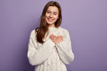 Wall Mural - Friendly pleased woman has heart filled with love and gratitude, keeps both hands on chest, has lovely sincere smile, wears oversized jumper, stands against purple background, sympatizes friend