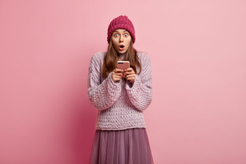 Sticker - Stupefied lovely woman holds cell phone, receives message with astonishing news, wears headgear, loose sweater and skirt, types feedback, isolated over pink background, surfes social network