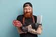 Isolated shot of pleased bearded male butcher crosses hands over chest, holds raw meet and cleaver, chopps on pieces, works in food industry, wears black clothes, has tattooed arms. Food concept