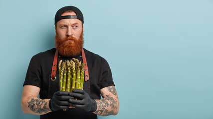 Wall Mural - Indoor shot of male chef holds spring vegetable, asparagus for preparing delicious salad, thinks which dish to cook, suggests healthy eating, has long ginger beard, wears black cap and t shirt