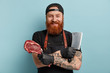 Headshot of cheerful friendly lookig butcher cuts meats in variety of piece, prepares for sale in retail environment, has pre sale processing of product holds ceaver and pork, wears cap, apron, gloves