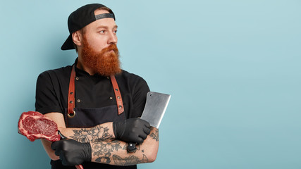 Sideways shot of handsome thoughtful male meat cutter prepares cuts of meat for selling, involved in idustrial processing, carries ceaver and raw meat, stands over blue background with free space