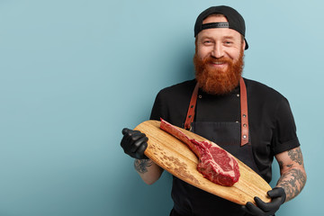 Wall Mural - Studio shot of cheerful bearded young man holds wooden board with piece of raw meat, wears black rubber gloves, going to cook supper in restaurant, works in food industry sphere. Cooking concept