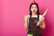 © sementsova321 - Portrait of young attractive woman in brown apron poses in studio, points thumb aside isolated on rose background. Astonished female stands with opened mouth, holds rolling pin. Culinary concept.