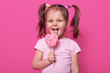 © sementsova321 - Happy little girl with striped lollypop in shape of heart in hand. Charming kid wears casual rose t shirt, with two funny pony tails. Cute child licks delicious candy isolated over pink background.