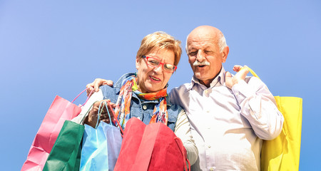 Senior couple shopping together with wife watching in husband bags - Elderly concept with mature man and woman having fun on sunny day - Happy retired people moments on vivid filter against blue sky