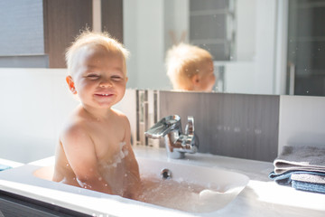 Canvas Print - Baby taking bath in sink. Child playing with foam and soap bubbles in sunny bathroom with window. Little boy bathing. Water fun for kids. Hygiene and skin care for children