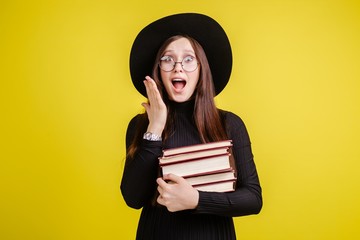 Beautiful girl with glasses and a black hat with her mouth open holding a stack of books in an isolated room on a yellow background