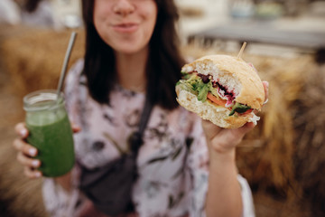 Stylish hipster girl holding delicious vegan burger and smoothie in glass jar in hands at street food festival, close up. Happy boho woman tasting burger with drink in summer street. Zero waste