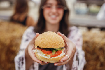 Wall Mural - Vegan burger closeup in boho girl hands. Stylish hipster girl in sunglasses eating delicious vegan burger at street food festival. Happy boho woman tasting and biting burger