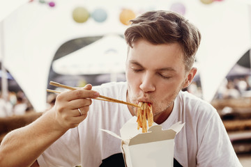 Stylish hipster man eating delicious wok noodles with vegetables from carton box with bamboo chopsticks. Asian Street food festival. Guy tasting and eating thai noodles in takeaway paper box