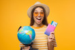 Excited female touristl holding globe in one hand and passport with tickets in other, ready to flight, isolated on yellow background