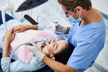 Wall Mural - Competent dentist in protective mask doing teeth cleaning for young lady