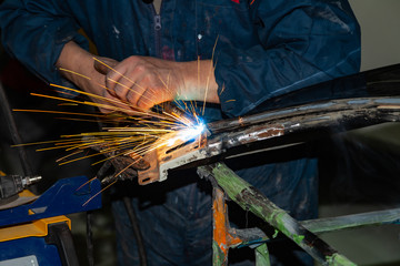 Wall Mural - Close-up view strong man is a welder in blue working overalls without gloves on arms, a metal product is welded with a welding machine in the garage workshop, blue and orange sparks fly to the sides