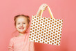 Closeup portrait of a cute little girl with hair buns over pink background. The child lifted the paper bag high, rejoicing at the shopping or gift.