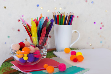 Child arts and craft work station with coloured pencils, colourful feathers, pom poms and paper on small round table with tablecloth