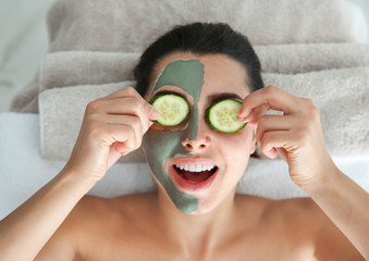 Poster - Pretty woman with clay mask on her face holding cucumber slices in spa salon, above view