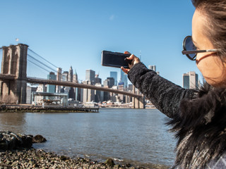 Young Woman tourist by the river in Dumbo taking pictures of Brooklyn Bridge and Cityscape of New York skyline.