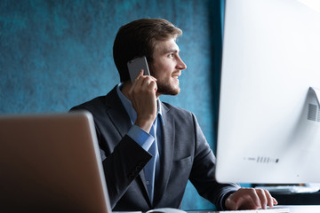 Portrait of handsome young male sitting at office desk with laptop computer and talking on mobile phone. Communication concept.