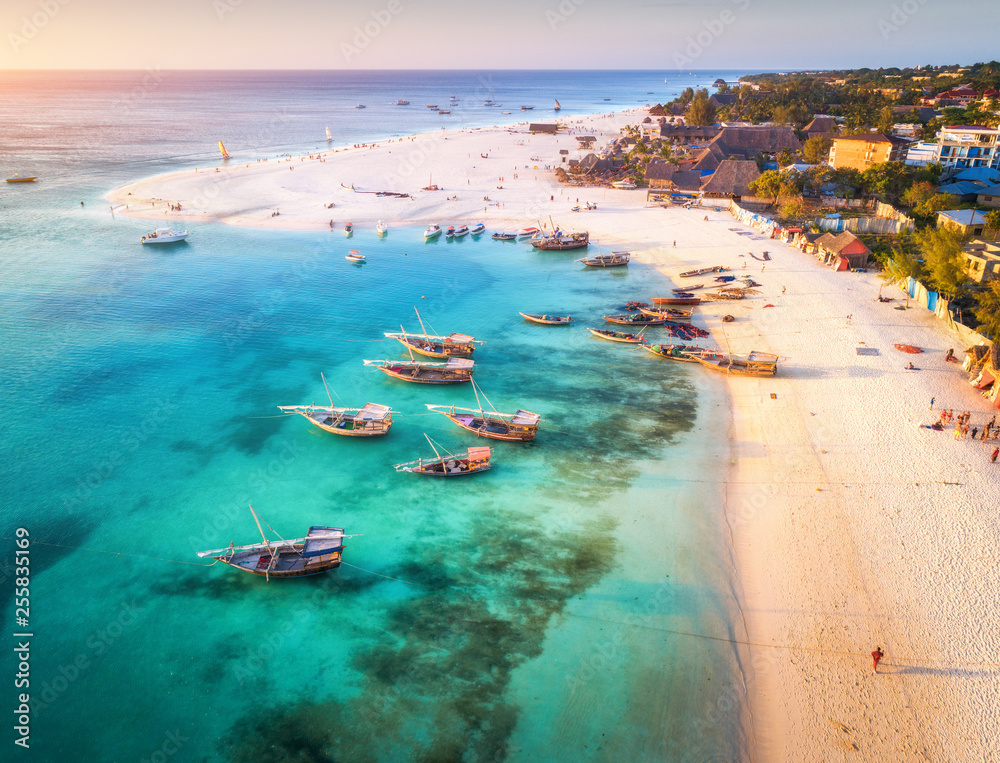 Aerial view of the fishing boats on tropical sea coast with sandy beach at sunset. Summer holiday on Indian Ocean, Zanzibar, Africa. Landscape with boat, buildings, transparent blue water. Top view - obrazy, fototapety, plakaty 