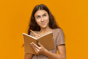 Creative teenage girl dressed in striped top holding diary and looking up with inspired facial expression searching for right words in her head while creating poem, making notes with pencil