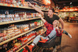 © estradaanton - Young couple in grocery store. Happy man and woman point on sauce shelf. She sit in trolley and has fun. Buying products in store.