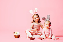 Portrait Of A Little Toddler Girl And A Baby With Bunny Ears And Easter Eggs Basket, On Pink