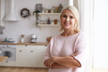 Attractive Middle Aged Housewife With Blonde Hair And Brown Eyes Posing Indoors In Her Modern Clean Stylish Kitchen Interior, Crossing Arms On Her Chest And Looking At Camera With Pleased Smile