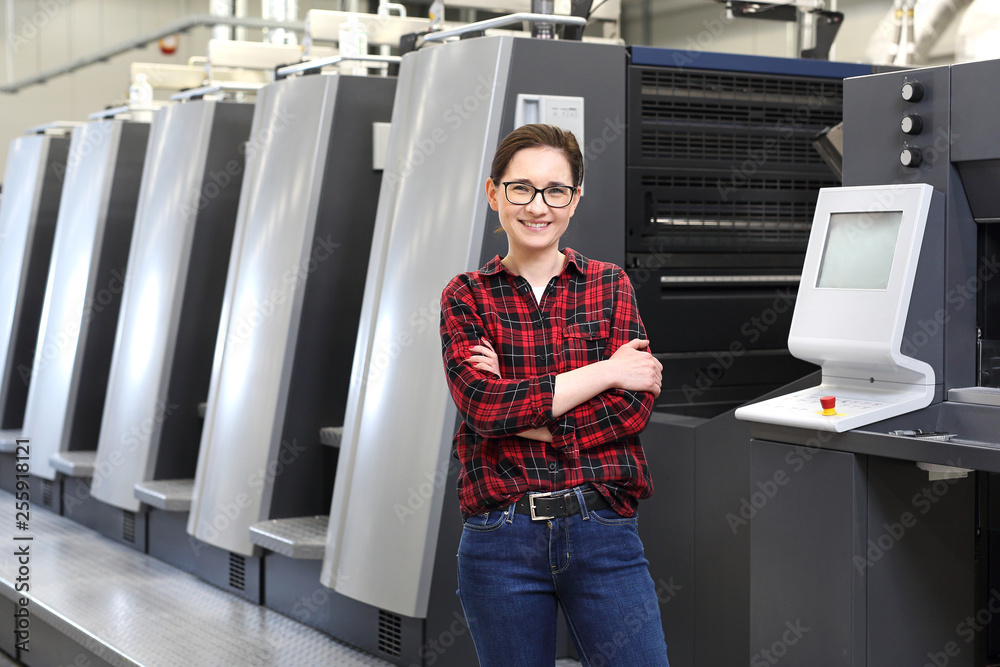 Praca w drukarni. Uśmiechnięta kobieta, pracownik drukarni stoi w hali produkcyjnej na tle nowoczesnych maszyn drukarskich - obrazy, fototapety, plakaty 