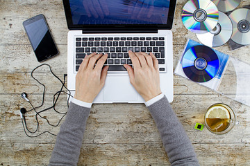 Young woman shooting from above working on laptop on wooden table listening to music cd with a cup of tea and headphone