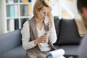 Wall Mural - Sad and depressed young woman wiping her tears and having glass of water while sitting on couch in psychologist office