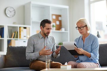 Wall Mural - Middle aged troubled man telling his story to mature female psychologist at individual session while both sitting on couch