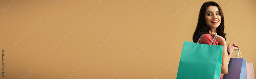 panoramic shot of smiling and beautiful woman holding shopping bags isolated on beige - obrazy, fototapety, plakaty 