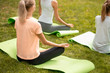 Young slim girl sits relaxing in the lotus position doing exercises on yoga mats with other girls on green grass in the park on a warm day.