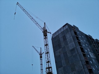 Two construction cranes and an unfinished house against the sky.