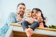 © Iryna - A low-angle view portrait of young happy family with a barefoot toddler girl sitting at kitchen table, saying 'Hi' and smiling. New home, happy healthy relationship concept.