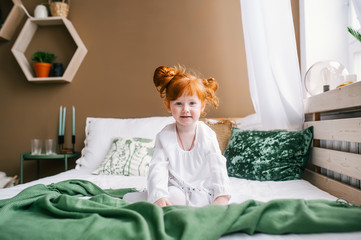 Cute happy redhead toddler girl sitting in bed at home