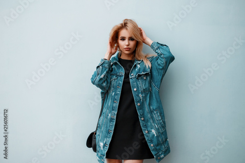 Cuadros en Lienzo American glamorous young blonde with a fashionable blue denim jacket in a stylish black dress with a leather handbag posing in a room near a vintage wall
