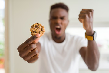 Sticker - Young african american man eating chocolate chips cookies annoyed and frustrated shouting with anger, crazy and yelling with raised hand, anger concept