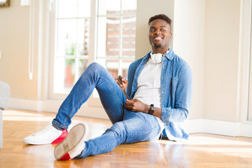 Poster - African american man wearing headphones listening to music sitting on the floor looking away to side with smile on face, natural expression. Laughing confident.