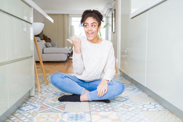 Beautiful young african american woman with afro hair sitting on the floor smiling with happy face looking and pointing to the side with thumb up.