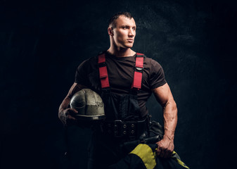 Wall Mural - Portrait of a brutal muscular fireman holding a helmet and jacket standing in the studio against a dark textured wall