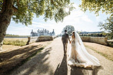 Elegant Newlywed Couple. Bride In A Luxurious Dress With A Long Bridal Veil. They Go To The Old Beautiful Castle In France