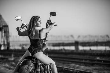 Red-haired woman in sexy lingerie in high heels sits on a motorcycle. Attractive red-haired girl sits on a motorcycle at sunset on the background of the industrial landscape