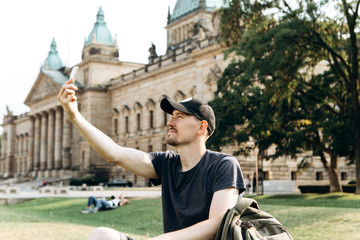 A young male tourist or student sits on the grass outside in Leipzig, Germany, talking on video or using a mobile phone to chat or take pictures.