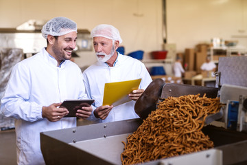 Supervisors in uniforms checking quality of food in food factory. Younger holding tablet while older holding folder with documents.