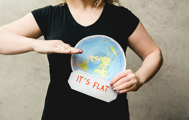  Flat Earther concept. Person who believes that Earth is flat disc. Anonymous woman holding flat Earth model in front of body with text: It`s flat. Isolated on gray background, studio shot. 